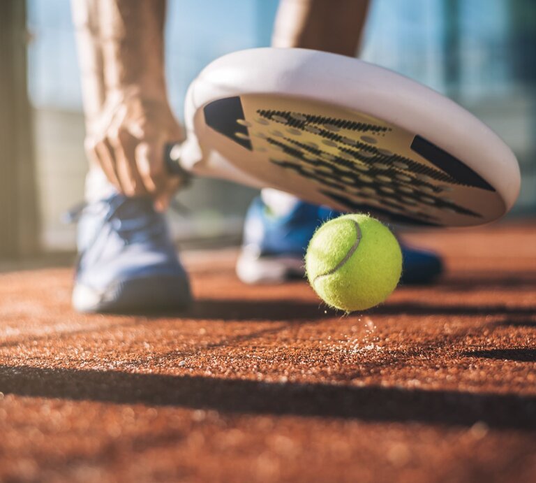 Eine Person in blauen Sportschuhen bereitet sich darauf vor, mit einem Padelschläger einen gelben Tennisball auf einem sandigen Platz zu schlagen. | © AdobeStock/DavidPrado
