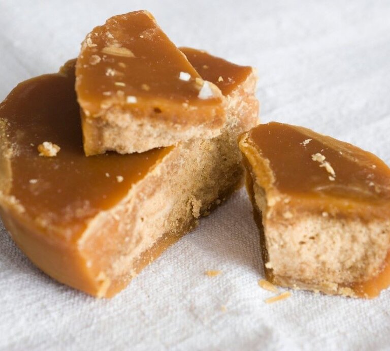 Das Bild zeigt Stücke von Jaggery, einem traditionellen, unraffinierten Zuckerprodukt, das oft in Südasien verwendet wird. Die goldbraune Färbung und die erkennbare Körnung weisen auf die natürliche Beschaffenheit hin. Der Jaggery scheint frisch gebrochen zu sein, was durch die unregelmäßigen Kanten und die sichtbaren Kristalle innerhalb des Zuckers deutlich wird. Die Textur sieht klebrig und feucht aus, typisch für hochwertigen Jaggery. Er liegt auf einer weißen Unterlage, die seine rustikale, natürliche Ästhetik betont. | © GettyImages / PhotoAlto/Laurence Mouton