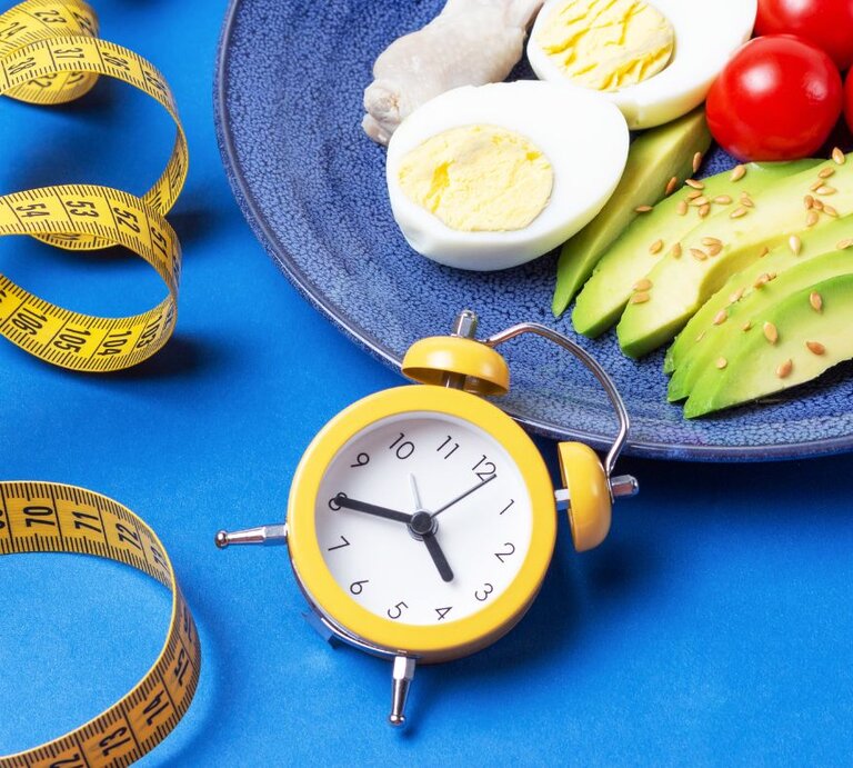 Ein Teller mit gesunden Lebensmitteln wie hartgekochten Eiern, Avocado-Scheiben mit Samen und Kirschtomaten, daneben ein gelber Wecker und ein Maßband auf einem blauen Hintergrund – Darstellung von Intervallfasten und gesunder Ernährung. | © Getty Images/ Tatiana via Canva.com
