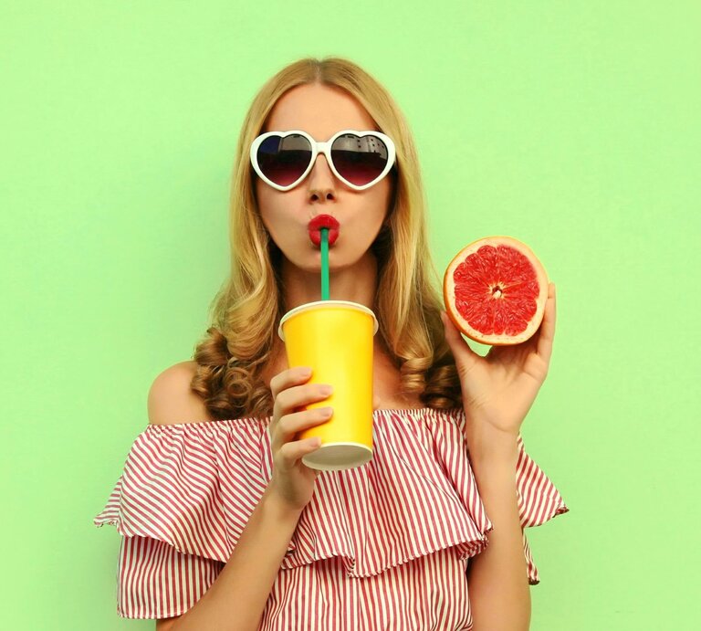 Eine blonde Frau vor grünem Hintergrund. Sie trägt eine Sonnenbrille, hält eine aufgeschnitte Grapefruit hoch und trinkt mit einem Strohhalm aus einem Becher. | © gettyimages/ Rohappy