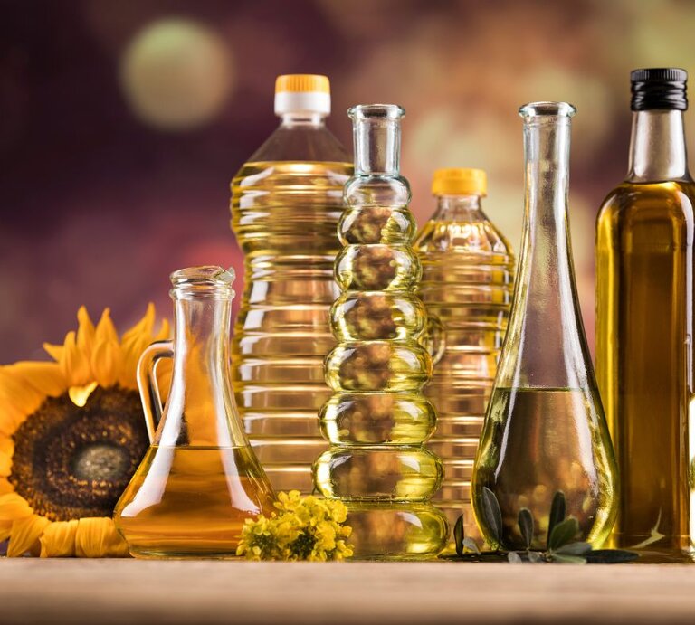 Verschiedene Flaschen mit Speiseöl aus Glas und Kunststoff stehen nebeneinander auf einem Tisch, darunter Sonnenblumenöl und Olivenöl; im Hintergrund sind eine Sonnenblume und warme, unscharfe Farben zu sehen. | © Adobe Stock/ Sebastian Duda