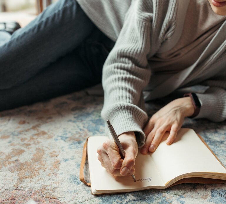 Eine Frau liegt auf dem Boden und schreibt in ein Tagebuch. | © Pexels/ Anastasia Shuraeva via Canva.com