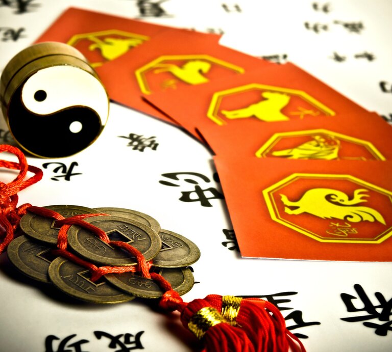  Eine detailreiche Darstellung chinesischer Astrologie: Im Vordergrund liegt ein traditioneller Glücksbringer mit Münzen und rotem Band. Daneben steht ein Yin-Yang-Symbol, umgeben von roten Karten mit gelben Illustrationen der chinesischen Tierkreiszeichen. Der Hintergrund zeigt schwarze chinesische Schriftzeichen auf weißem Papier. Das Bild vermittelt eine Atmosphäre von Tradition, Weisheit und fernöstlicher Kultur. | © Adobe Stock/ starblue