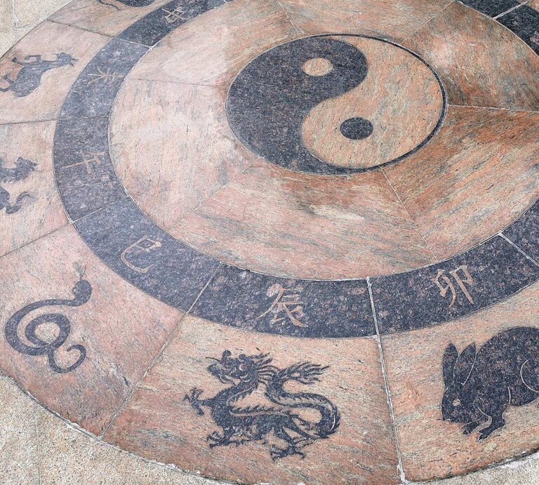 Ein runder, steinerner Boden mit gravierten Symbolen der chinesischen Tierkreiszeichen, darunter Drache, Schlange, Hase und Pferd. In der Mitte ist das Yin-Yang-Symbol abgebildet, das die Balance von Gegensätzen in der chinesischen Philosophie symbolisiert. | © Getty Images Signature/ gionnixxx via Canva.com
