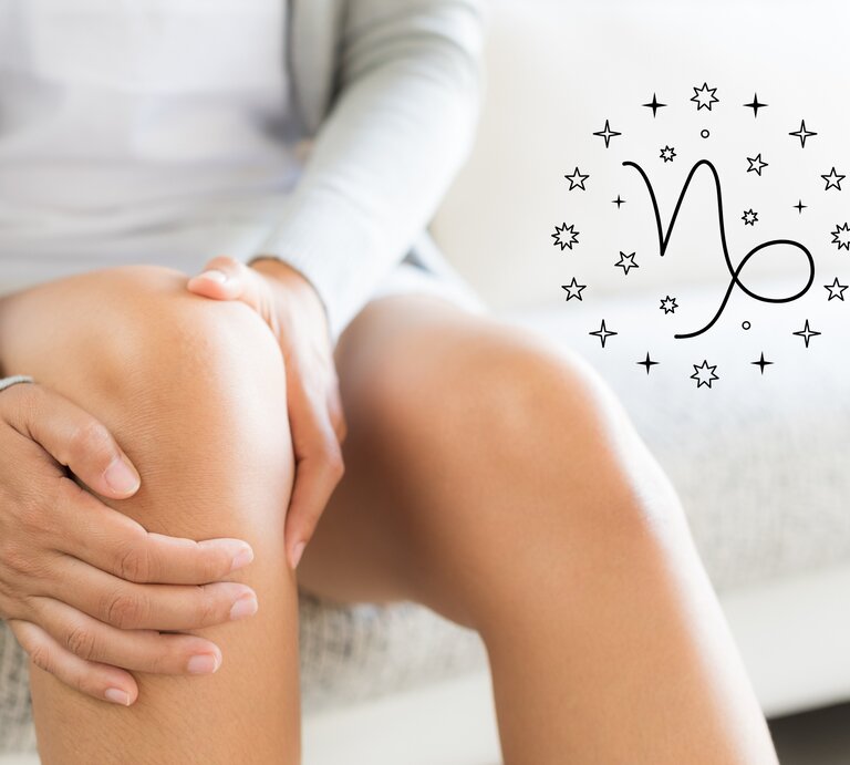 Das Bild zeigt die Hände einer Person, die sanft das rechte Knie umfasst. Die Person sitzt auf einem Sofa und trägt eine hellgraue Strickjacke und ein weißes Kleid. Rechts neben dem Knie ist das astrologische Symbol für Steinbock (Capricorn) abgebildet, umgeben von kleinen Sternen. Der Hintergrund ist hell und vermittelt eine ruhige und entspannte Atmosphäre. | © Getty Images Pro/ spukkato und warmjuly via Canva.com