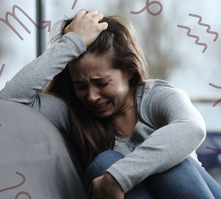 Eine junge Frau sitzt weinend und verzweifelt auf einem Sofa. Sie greift sich mit einer Hand in die Haare und wirkt sehr emotional und gestresst. Um sie herum sind verschiedene astrologische Symbole in einem dezenten Braunton zu sehen. Der Hintergrund ist unscharf und zeigt Bäume und ein Fenster, wodurch eine bedrückende Stimmung erzeugt wird. | © Adobe Stock/ Antonioguillem und Keronyart's Images/ Be keronyart via Canva.com [M]