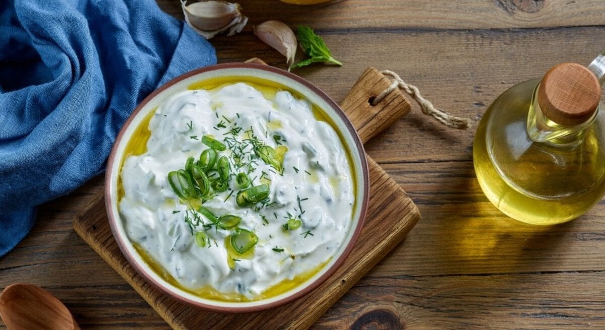 Ein farbenfrohes Bild zeigt eine Schale Çaçık, eine türkische Joghurtspeise, garniert mit geschnittenen Frühlingszwiebeln und Dill, auf einem Holzbrett. Neben der Schale befinden sich eine Zitrone, einige Knoblauchzehen, ein Bund frischer Minze und eine kleine Flasche Olivenöl mit einem Holzkorken. Eine blaue Stoffserviette liegt stilvoll gefaltet neben der Schale. Alle Elemente sind auf einer rustikalen Holzoberfläche arrangiert, die Wärme und Natürlichkeit ausstrahlt.
