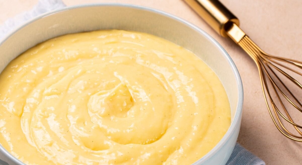 Low Carb Nachtisch: Cremiger Vanillepudding in einer hellblauen Schale auf einem hellblauen Tuch, daneben Vanilleschoten und ein goldener Schneebesen.