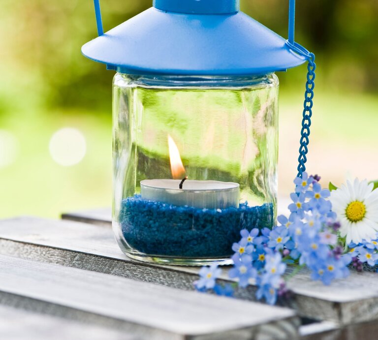 Ein Windlicht mit blauer Metallhaube und brennender Kerze steht auf einem Holztisch, umgeben von blauen Blüten. | © Adobe Stock/DoraZett