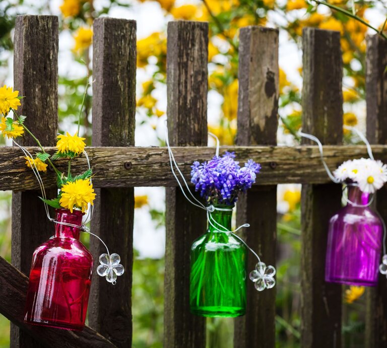 Drei bunte Glasflaschen mit gelben, blauen und weißen Blumen hängen dekorativ an einem alten Holzzaun im blühenden Garten. | © Adobe Stock/fotoknips