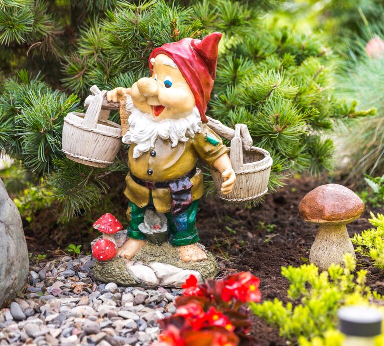 Ein bunter Gartenzwerg mit roter Mütze und weißen Bart trägt zwei Holzeimer und steht lachend zwischen grünen Pflanzen und Pilzen. | © Adobe Stock/Mariakray