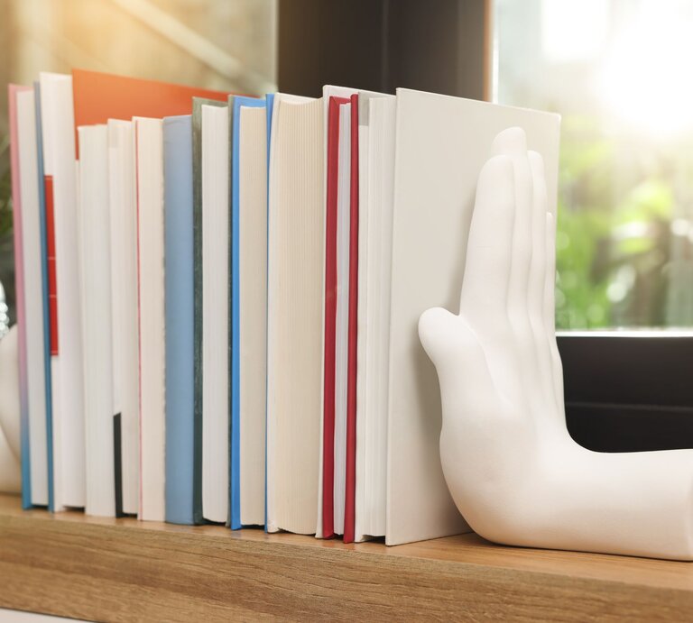 Eine Reihe bunter Bücher steht auf einem Holzregal, gestützt von weißen Buchstützen in Form einer Hand vor einem sonnigen Fenster. | © Adobe Stock/New Africa