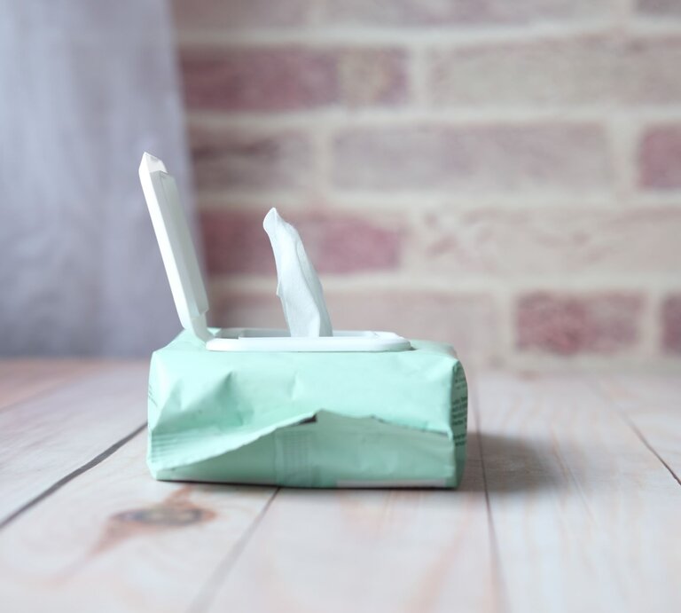 Eine geöffnete Packung Feuchttücher in mintgrüner Verpackung steht auf einem hellen Holztisch vor einer Backsteinwand. | © Adobe Stock/Towfiqu Barbhuiya