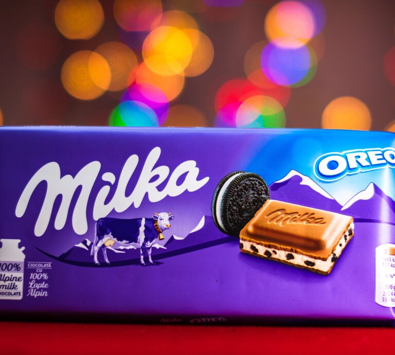 Eine Tafel Milka-Oreo-Schokolade in lilafarbener Verpackung mit Alpenmotiv und Milka-Kuh, vor buntem, unscharfem Lichterhintergrund. | © Adobe Stock/Vlad Ispas
