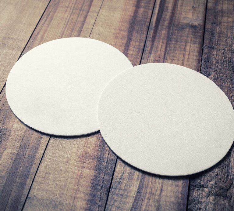 Schlichte, unbedruckte Bierdeckel auf gealtertem Holzuntergrund mit sichtbarer Maserung und warmem Farbton. | © Adobe Stock/Veresovich
