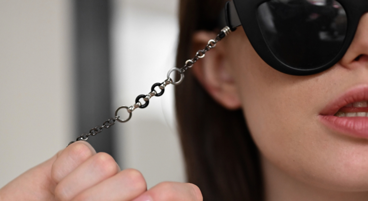 Junge Frau mit braunem Haar und schwarzer Cat-Eye-Sonnenbrille mit Kette, schwarze Sonnenbrille im Sandstrand vor Meer, schwarze Lesebrille und Schlüssel auf Notizbuch neben Tasse.
