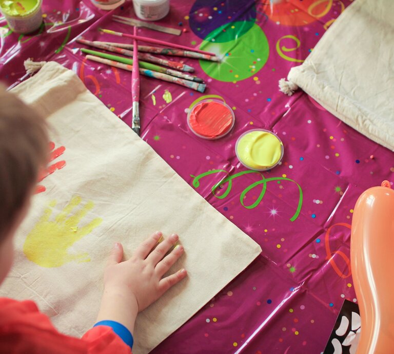 Ein Kind drückt seine Hand in gelber Farbe auf eine Stofftasche, umgeben von Pinseln und Farbtöpfen auf pinkem Partytuch. | © Adobe Stock/MStock