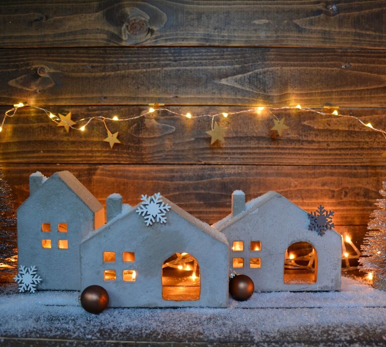 Kleine, weiße Häuser aus Ton mit Schneeflocken-Deko und Lichterkette stehen auf Kunstschnee vor einer Holzwand, umgeben von glitzernden Tannen. | © Adobe Stock/S.H.exclusiv