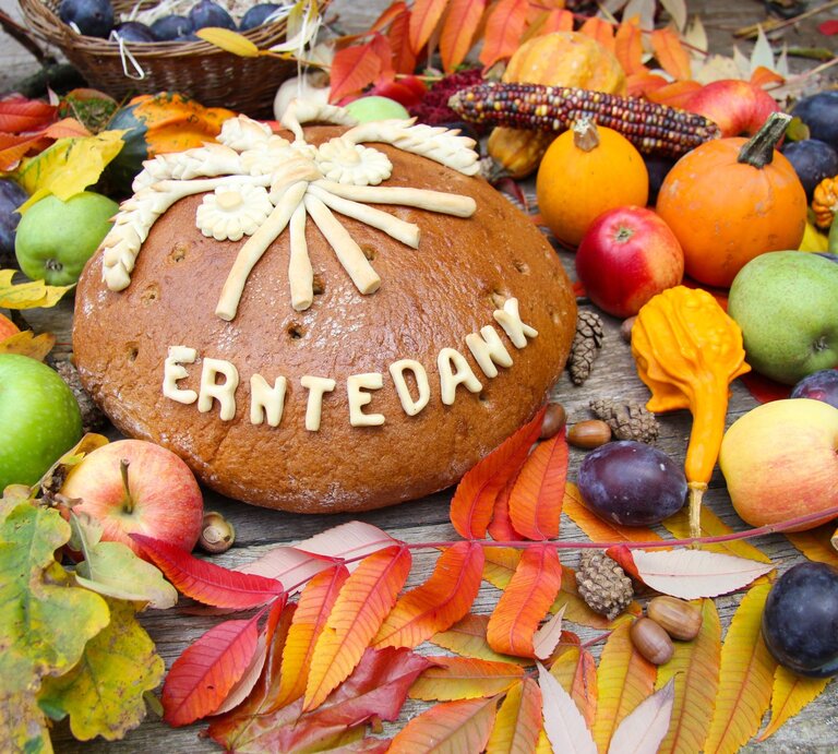 Großer Laib Erntedankbrot mit Verzierung liegt umgeben von Äpfeln, Kürbissen, Maiskolben und buntem Herbstlaub. | © Adobe Stock/ChristArt