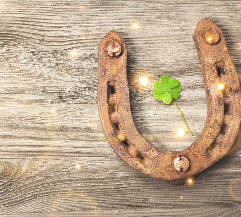Rostiges Hufeisen auf hellem Holzbrett mit grünem Kleeblatt in der Mitte und sanft funkelnden Lichtern. | © Adobe Stock/BillionPhotos.com