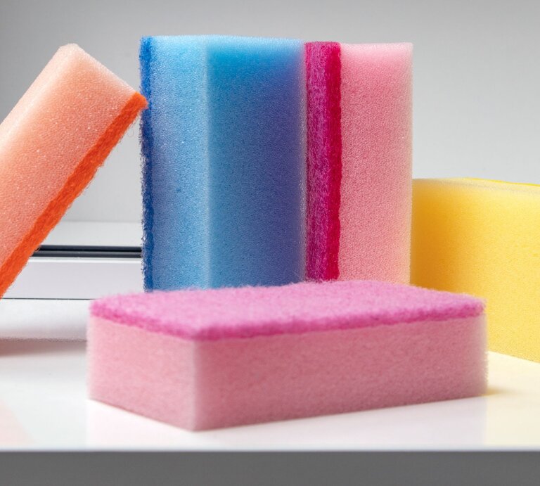 Bunte Reinigungsschwämme in Gelb, Blau, Rosa und Orange stehen aufrecht und liegen auf einem weißen Tisch. | © Adobe Stock/Alex Shin