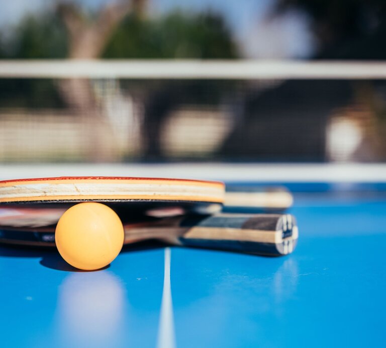 Zwei Tischtennisschläger und ein Ball liegen auf einem blauen Tisch vor unscharfem Hintergrund im Freien. | © Adobe Stock/antoniosantosc