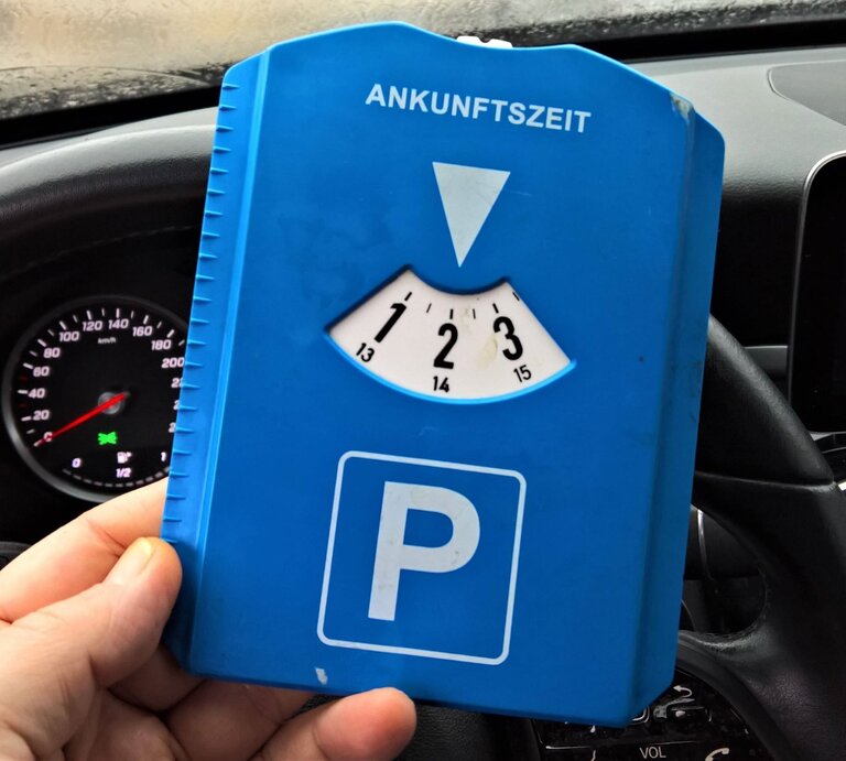 Eine Hand hält eine blaue Parkscheibe mit der Ankunftszeit auf 14 Uhr vor einem Autolenkrad und einem Tacho. | © IMAGO / Manfred Segerer