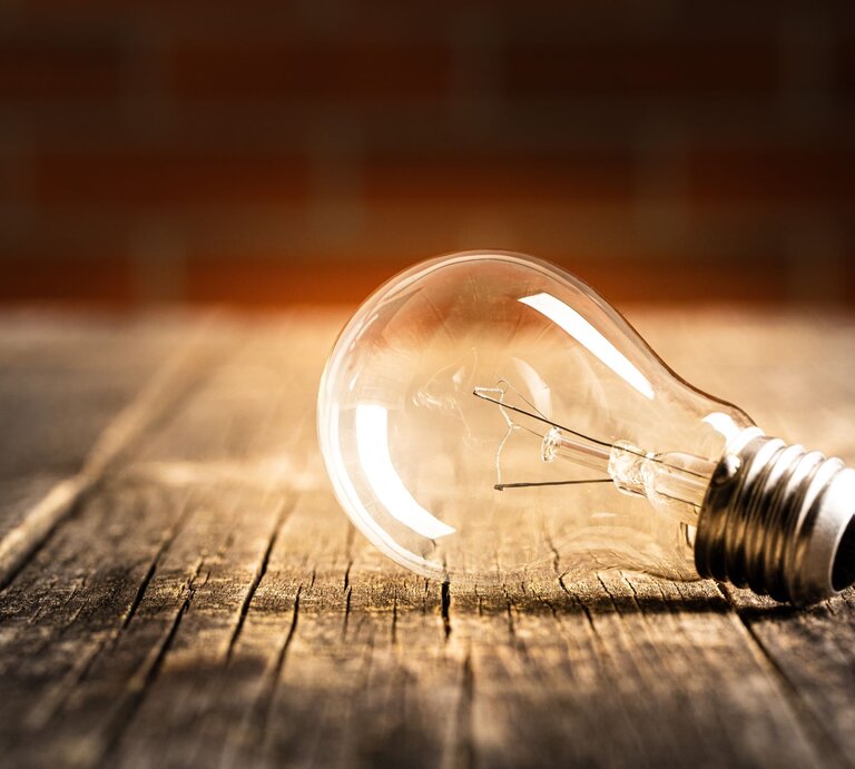 Glühbirne liegt unbeleuchtet auf einer rustikalen Holzoberfläche, beleuchtet von warmem Licht. | © Adobe Stock/Jiri Hera
