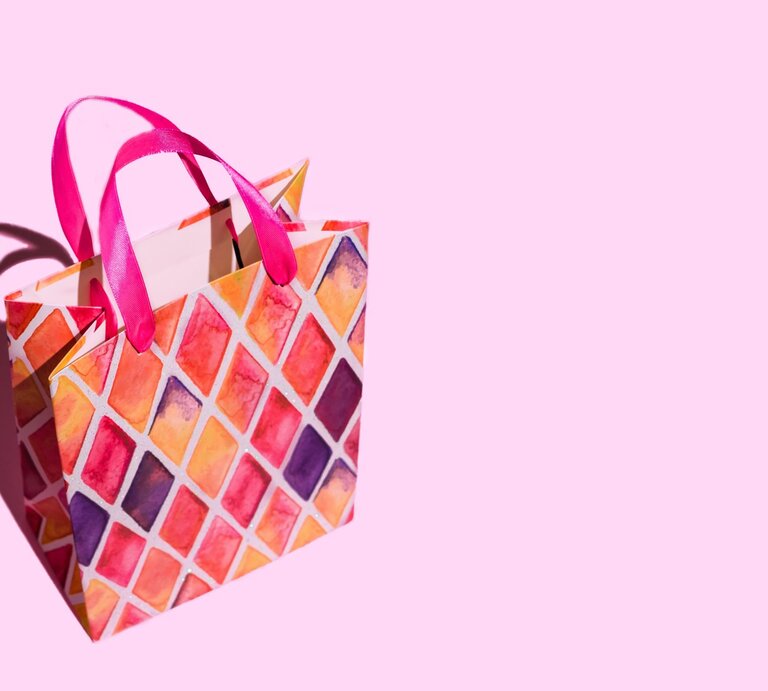 Bunte Geschenktüte mit pinken Griffen und rautenförmigem Muster in Rot-, Orange- und Lilatönen auf rosa Hintergrund. | © Getty Images/Iryna Veklich