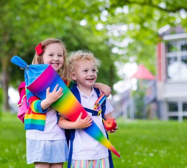 Zwei fröhliche Kinder, eines mit Locken und eines mit Haarspange, halten eine bunte Schultüte im Grünen vor einem Schulgebäude. | © Adobe Stock/famveldman