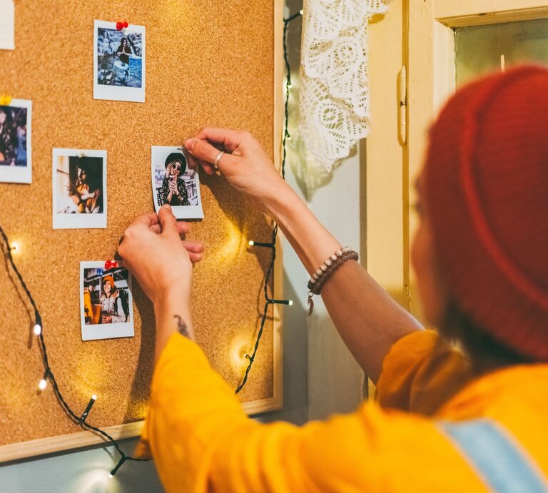 Junge Frau mit roter Mütze und gelbem Pullover pinnt Polaroid-Foto an eine Pinnwand aus Kork mit Lichterkette. | © Getty Images/martin-dm