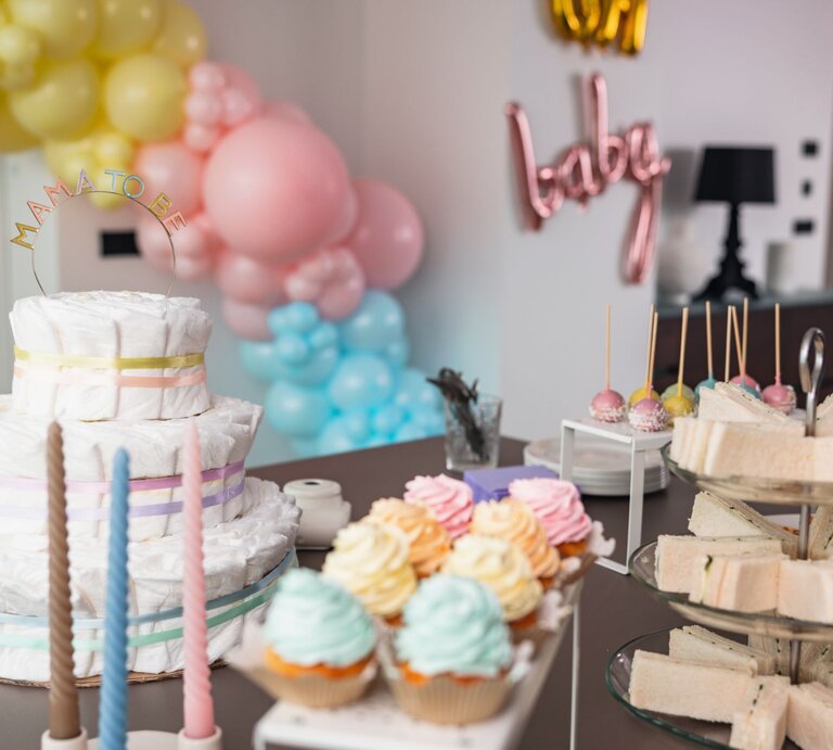 Ein Tisch mit einer Windeltorte, bunten Cupcakes, Sandwiches und Cake-Pops bei einer Babyparty mit Luftballon-Dekoration. | © Getty Images/AzmanJaka