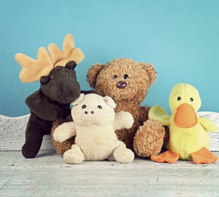Vier bunte Stofftiere, darunter ein Elch, ein Teddy, ein Schweinchen und eine Ente, sitzen vor einer blauen Wand auf hellem Holzboden. | © Adobe Stock/ARTFULLY-79