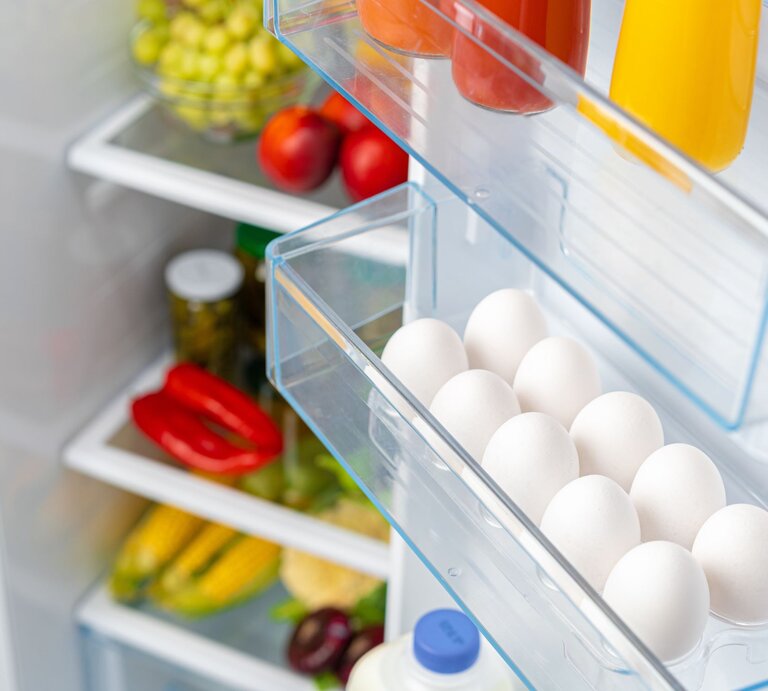 Ein geöffneter Kühlschrank zeigt Eier, Säfte und frisches Gemüse wie Paprika und Mais in den Regalen. | © Adobe Stock/fotofabrika