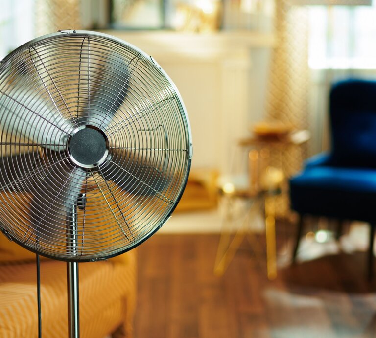 Ein silberner Standventilator steht in einem gemütlich eingerichteten Wohnzimmer mit blauen Sesseln und Holzboden. | © Adobe Stock/Alliance