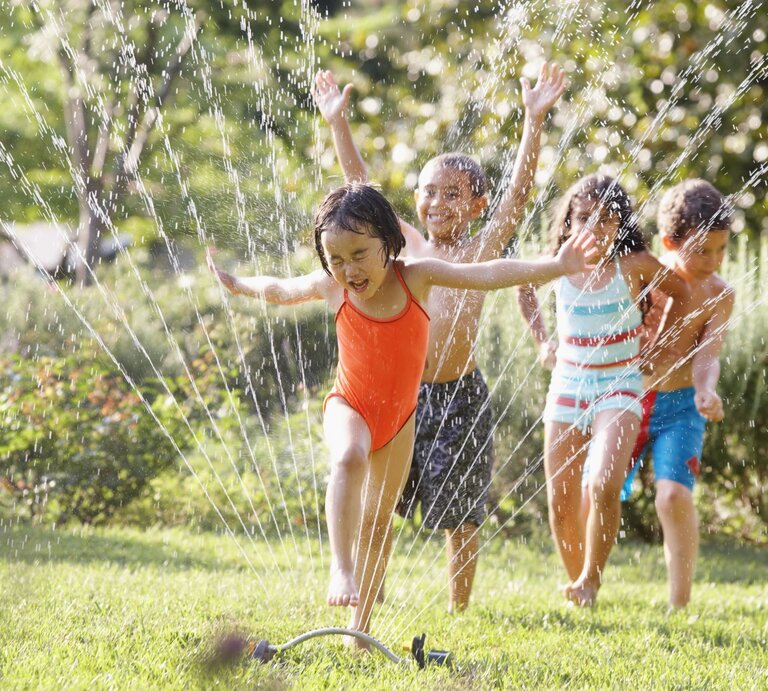 Vier Kinder in bunter Badebekleidung rennen lachend durch Wasserstrahlen eines Rasensprengers auf einer grünen Wiese an einem sonnigen Tag. | © Getty Images/Ariel Skelley
