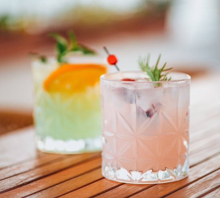 Zwei erfrischende Cocktails in Kristallgläsern auf einem Holztisch, der vordere rosa mit Rosmarin und Beeren, der hintere grün mit Orangenscheibe und Minze. | © Getty Images/Dudits