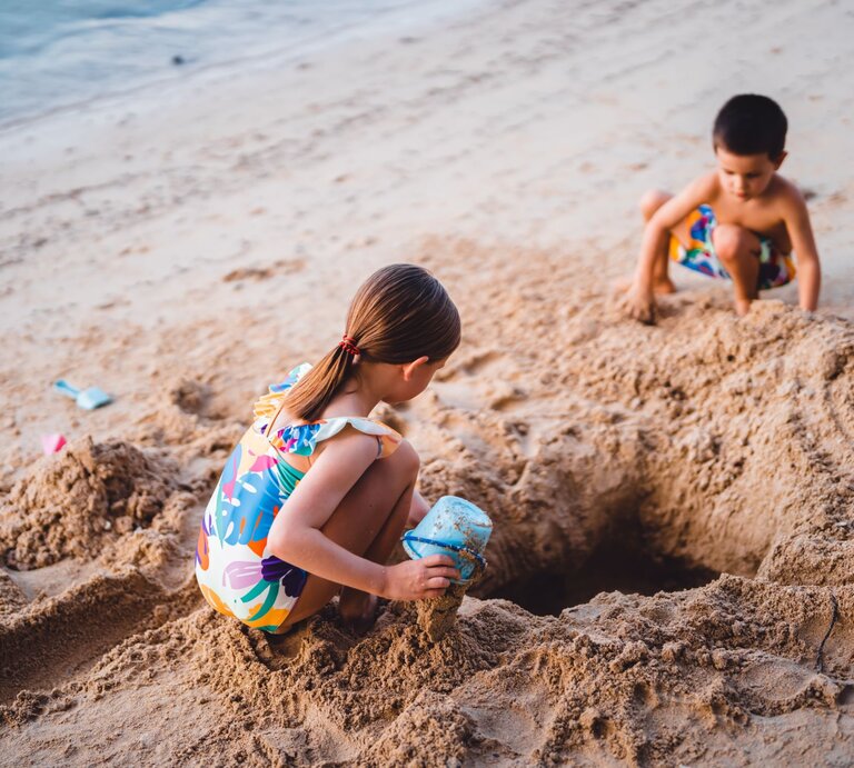 Zwei junge Kinder in farbenfroher Strandkleidung buddeln im Sand an einem leeren Strand ein Loch. | © Getty Images/Yulia Grossman