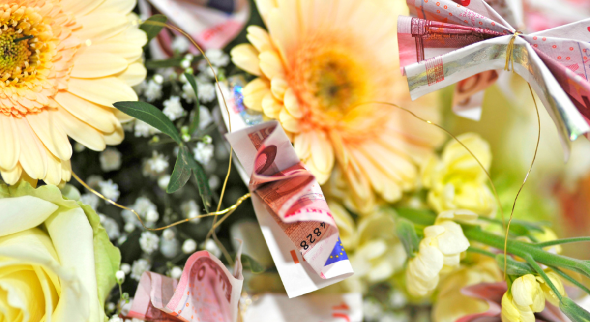 Brautpaar küsst sich unter dem Schleier, daneben ein Geldgeschenk mit Schleife und ein Blumenstrauß aus Geldscheinen und Gerbera.