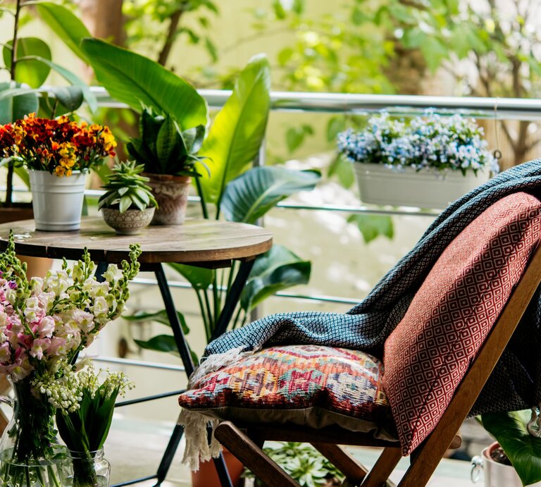 Ein gemütlicher Balkon mit Holztisch, bunten Blumentöpfen, gemustertem Kissen und Decke auf Klappstuhl vor üppigem Grün. | © Shutterstock/fornStudio