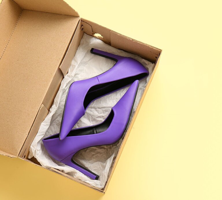 Ein Paar lila High Heels mit spitzen Absätzen liegt in einer geöffneten Pappschachtel auf gelbem Hintergrund. | © Adobe Stock/Pixel-Shot
