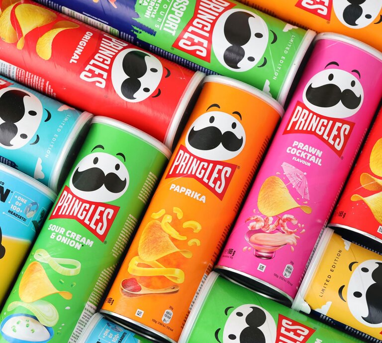 Bunte Pringles-Dosen in verschiedenen Geschmacksrichtungen wie Paprika, Sour Cream und Prawn Cocktail liegen nebeneinander aufgereiht. | © Adobe Stock/mehaniq41
