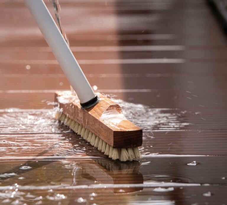 Grobe Bürste mit Holzgriff schrubbt unter fließendem Wasser eine nasse, braune Terrassendiele bei Sonnenlicht. | © Shutterstock/Tamara B