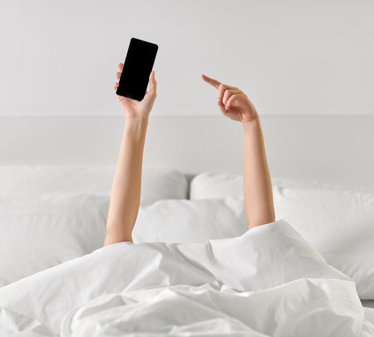 Zwei Arme strecken sich aus einer weißen Bettdecke, einer hält ein Smartphone, der andere zeigt mit dem Finger nach oben. | © AdobeStock/Syda Productions