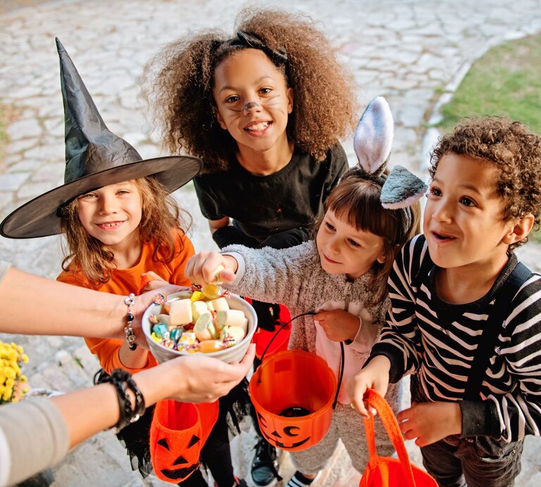 Vier fröhliche Kinder in Halloween-Kostümen halten orangefarbene Eimer und nehmen Süßigkeiten aus einer Schale entgegen. | © AdobeStock/Tijana