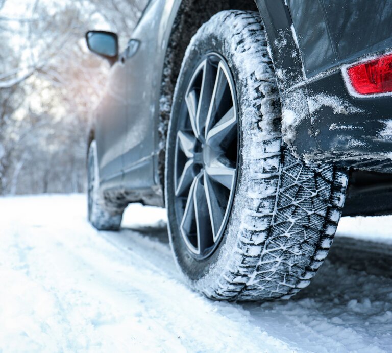 Ein dunkles Auto mit Schneereifen fährt auf einer verschneiten Straße durch einen winterlichen Wald mit kahlen Bäumen. | © AdobeStock/New Africa