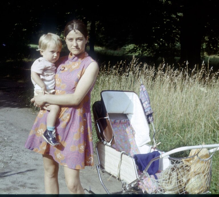 Mutter in lila Kleid mit Kind auf dem Arm, steht neben einem weißen Kinderwagen, im Hintergrund hohes Gras. | © Getty Images/dm909