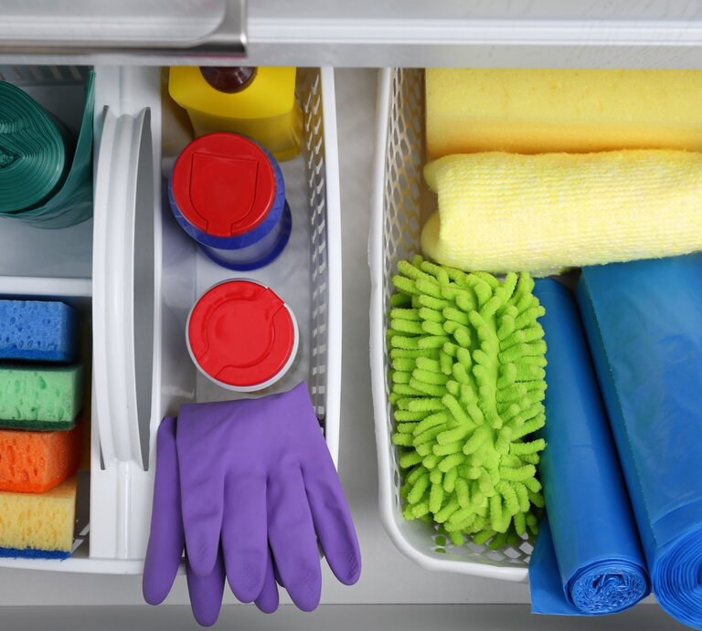 Geordnete Reinigungsmittel in weißen Körben: Schwämme, Müllbeutel, violette Handschuhe und Mikrofasertücher in Gelb und Grün. | © Adobe Stock/New Africa