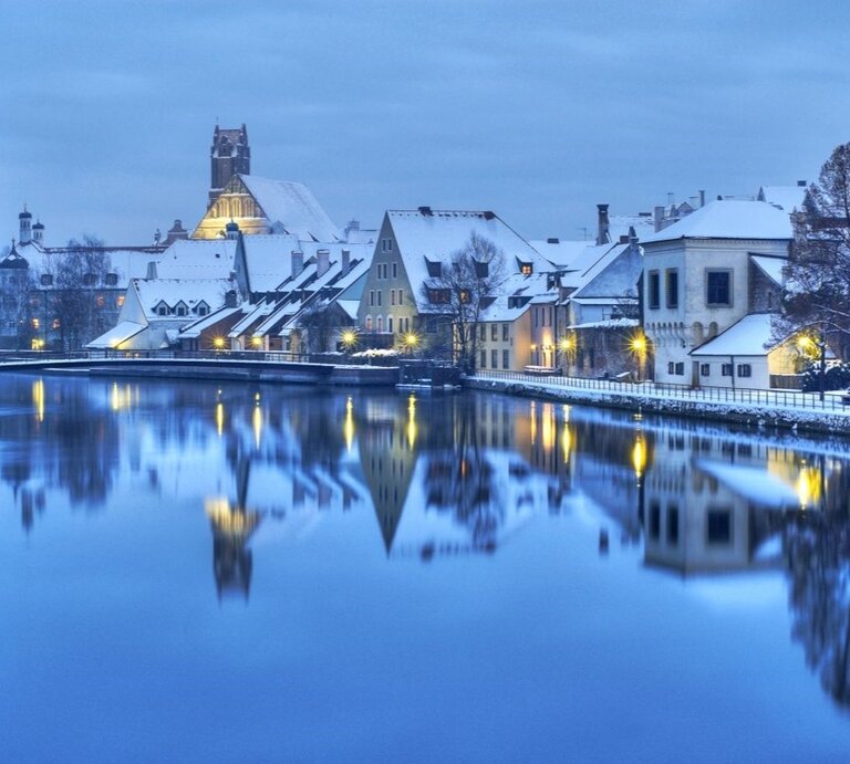 Eine verschneite Altstadt am Abend. Die Lichter spiegeln sich im Fluss. | © Shutterstock/Boris Stroujko