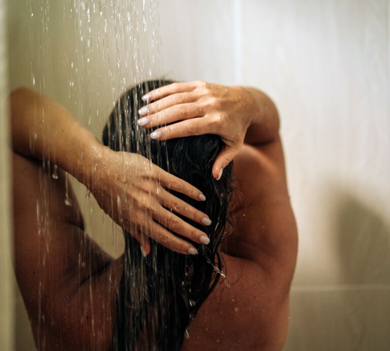 Eine Frau mit nassen Haaren steht in der Dusche und hält sich die Hände über den Kopf, während Wasser auf sie herabströmt. | © GettyImages/Fiordaliso